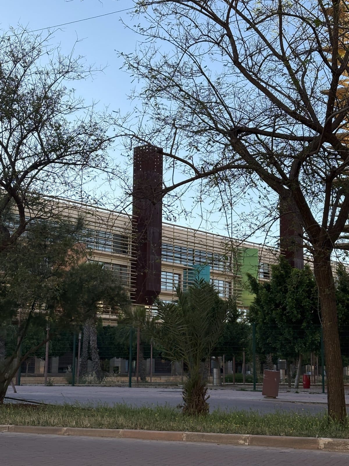 1337 Khouribga by day: a long building behind louvres and trees, with a perforated rust-red tower